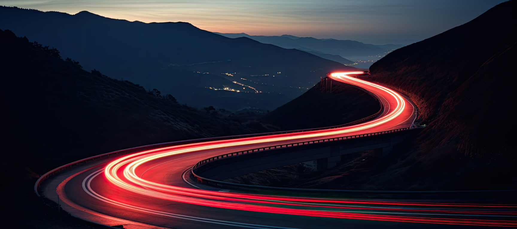 Winding mountain road with vehicle light trails at dusk, illustrating long automotive travel times and delays that private jet travel can help reduce.